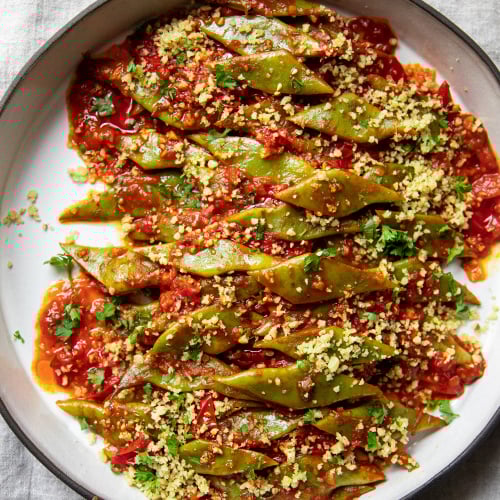 An overhead image of braised Romano beans in a round ceramic dish. The beans are topped with ground walnuts and parsley.