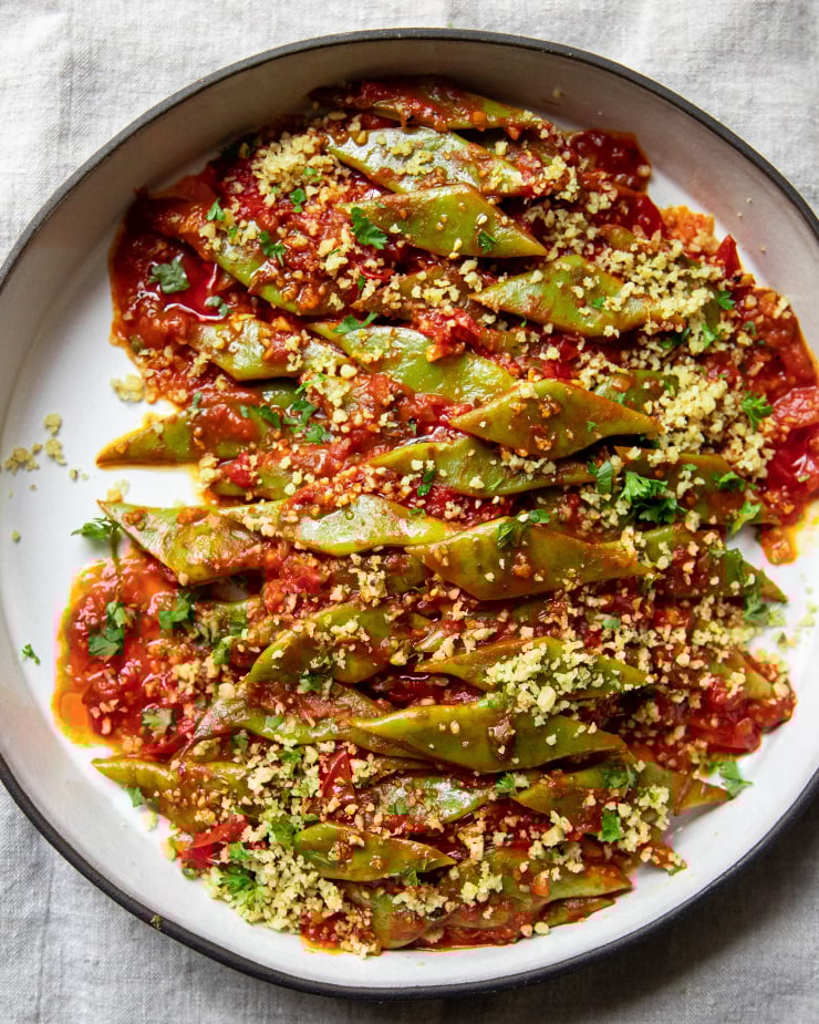 An overhead image of braised Romano beans in a round ceramic dish. The beans are topped with ground walnuts and parsley.