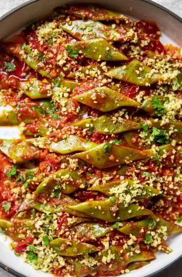 An overhead image of braised Romano beans in a round ceramic dish. The beans are topped with ground walnuts and parsley.