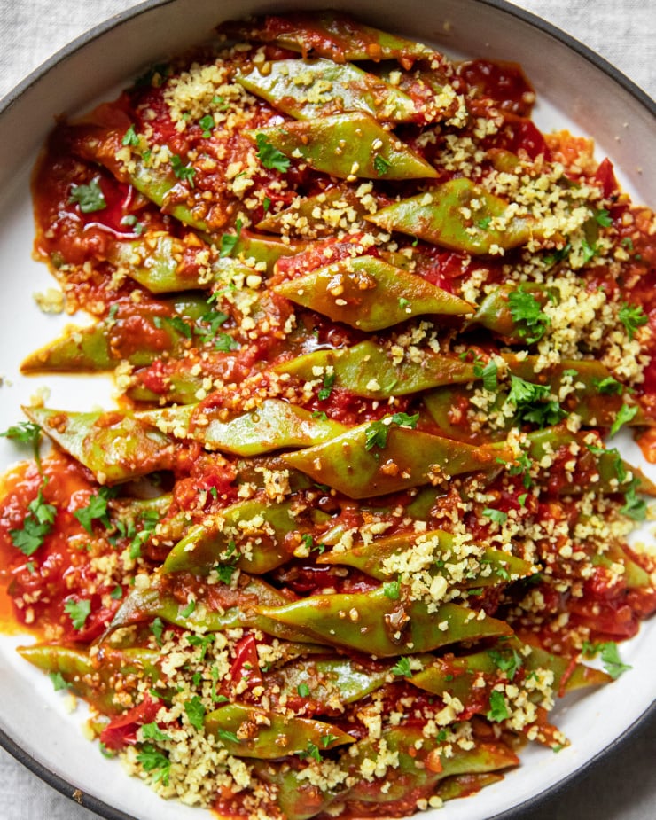 An overhead image of braised Romano beans in a round ceramic dish. The beans are topped with ground walnuts and parsley.