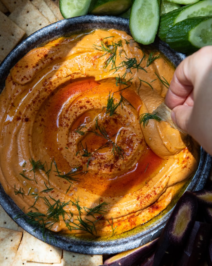 An overhead image of a creamy orange dip in a dark blue shallow bowl. The dip is surrounded by cut vegetables and crackers. A hand is seen swooping a cracker through the dip.