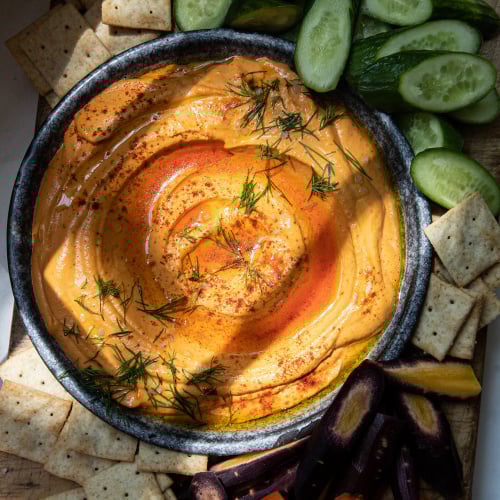 An overhead image of a creamy orange dip in a dark blue shallow bowl. The dip is surrounded by cut vegetables and crackers.