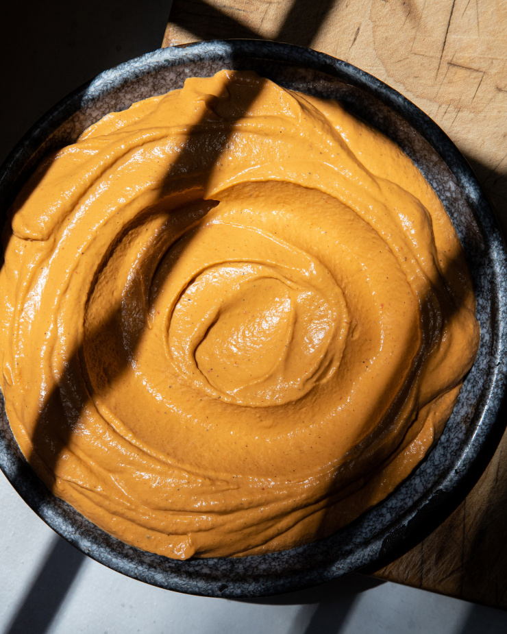 An overhead shot of a creamy orange-hued vegan red pepper dip in a shallot, dark blue plate. The dip has been swooped with a spoon to create swirls on top.