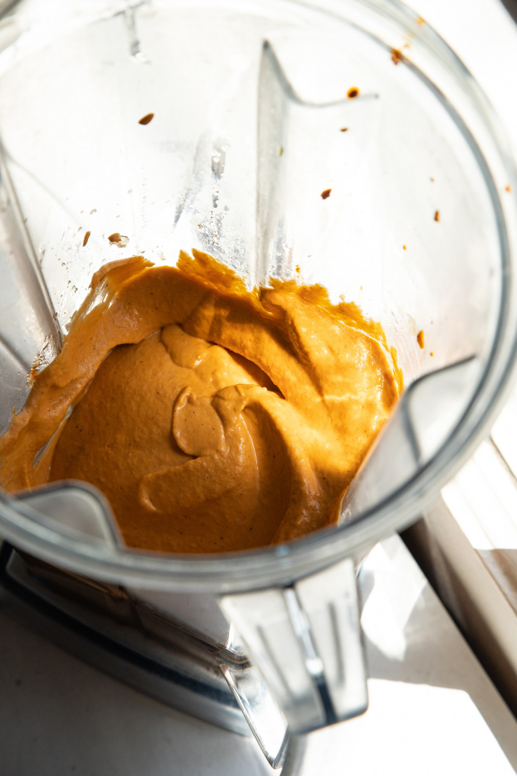 A 3/4 angle shot of a blended up orange-hued vegan red pepper dip in a blender pitcher.