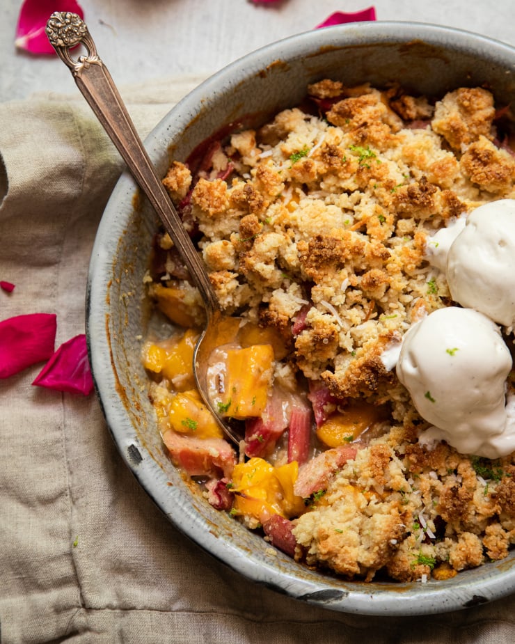 An up close, overhead shot of a rhubarb mango crumble in a grey enamelware, round dish. It has a crisp, beige cookie-like topping. The crumble has two melt-y scoops of ice cream on top.