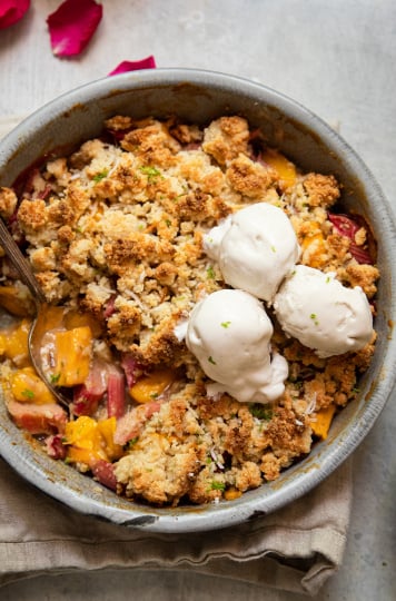 An up close, overhead shot of a rhubarb mango crumble in a grey enamelware, round dish. It has a crisp, beige cookie-like topping. The crumble has two melt-y scoops of ice cream on top.