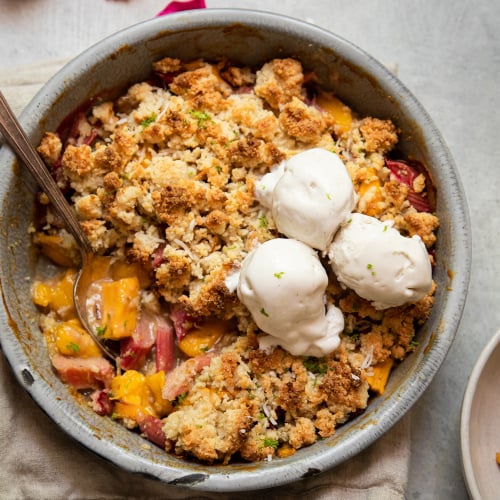 An up close, overhead shot of a rhubarb mango crumble in a grey enamelware, round dish. It has a crisp, beige cookie-like topping. The crumble has two melt-y scoops of ice cream on top.