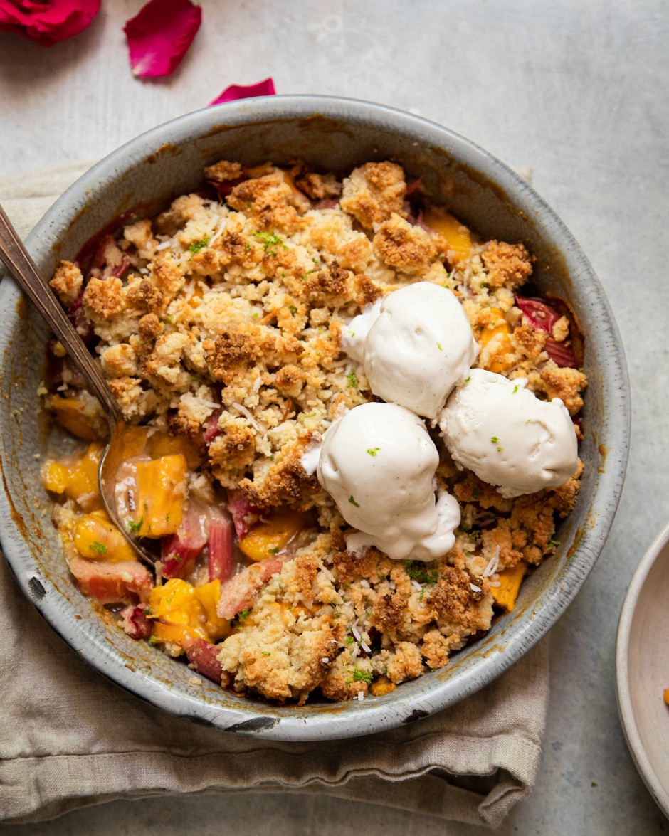 An up close, overhead shot of a rhubarb mango crumble in a grey enamelware, round dish. It has a crisp, beige cookie-like topping. The crumble has two melt-y scoops of ice cream on top.