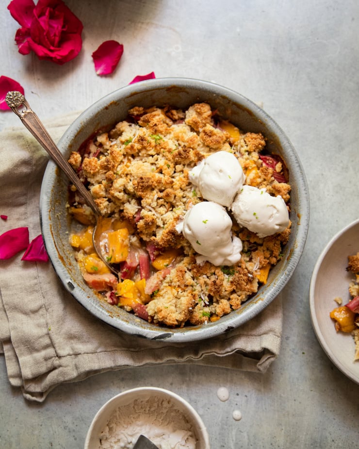 An overhead shot of a rhubarb mango crumble in a grey enamelware, round dish. It has a crisp, beige cookie-like topping. The crumble has two melt-y scoops of ice cream on top.