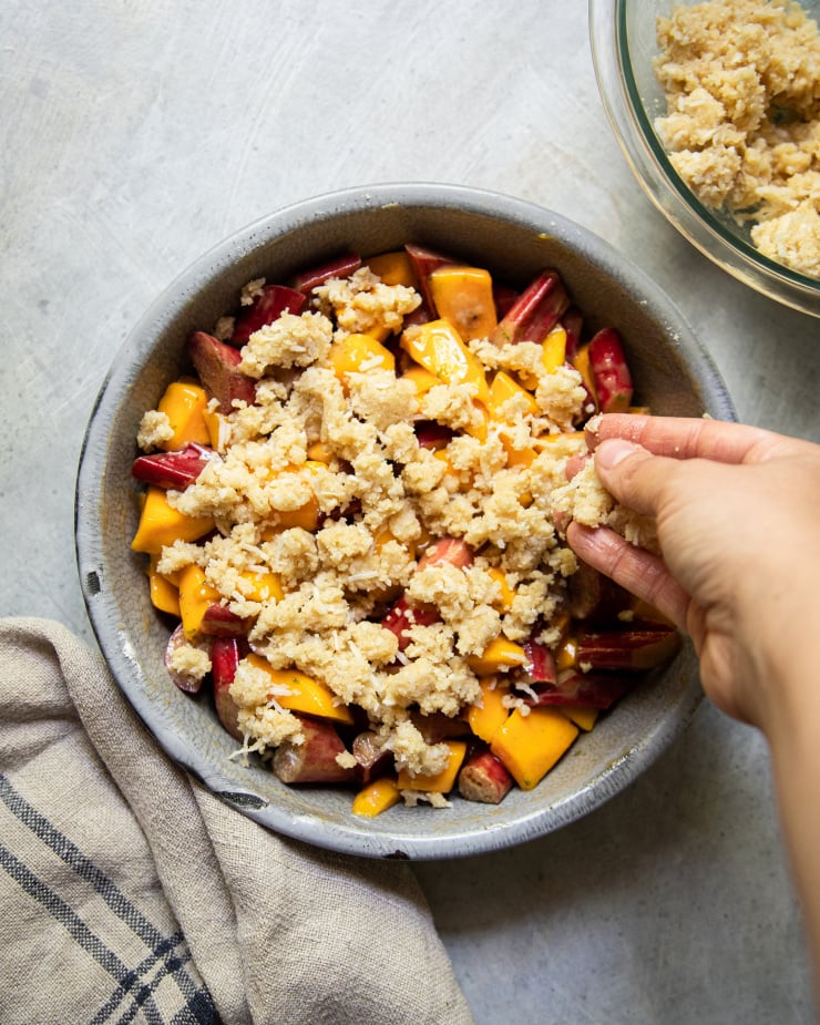 A hand is sprinkling crumble topping onto a dish of chopped fruit. This image is shot from overhead on a grey background.