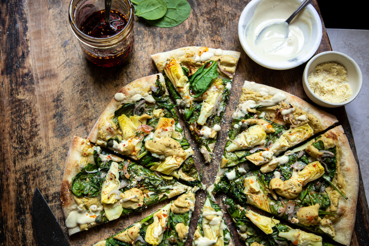 Overhead: up close image of a sliced vegan spinach artichoke pizza. The blade of a knife is nearby. Pizza is shown on a worn wood background.