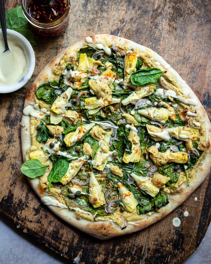 Overhead: image of a vegan spinach artichoke pizza. Pizza is shown on a worn wood background. A small bowl of creamy sauce is shown nearby.