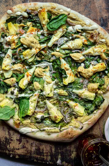 Overhead: up close image of a vegan spinach artichoke pizza. Pizza is shown on a worn wood background. A small bowl of creamy sauce is shown nearby.