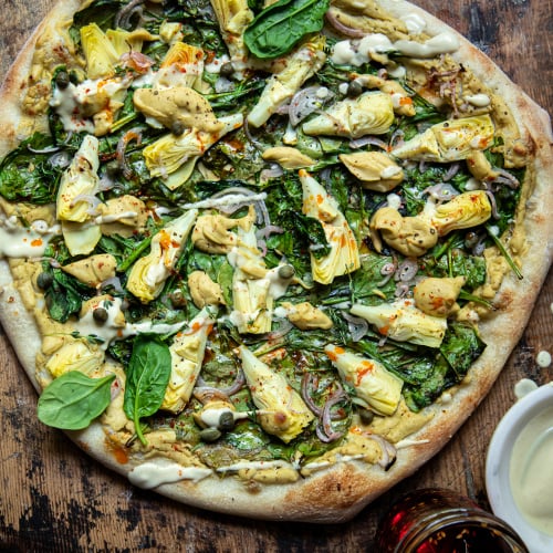 Overhead: up close image of a vegan spinach artichoke pizza. Pizza is shown on a worn wood background. A small bowl of creamy sauce is shown nearby.