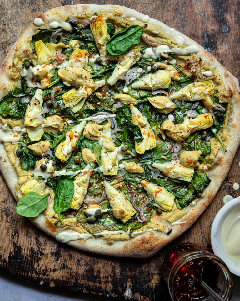 Overhead: up close image of a vegan spinach artichoke pizza. Pizza is shown on a worn wood background. A small bowl of creamy sauce is shown nearby.