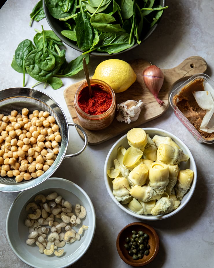An overhead shot of ingredients on a mottled grey background. There are: chickpeas, cashews, artichoke hearts, ground chillies, miso, capers, a lemon, garlic, a shallot, and baby spinach.