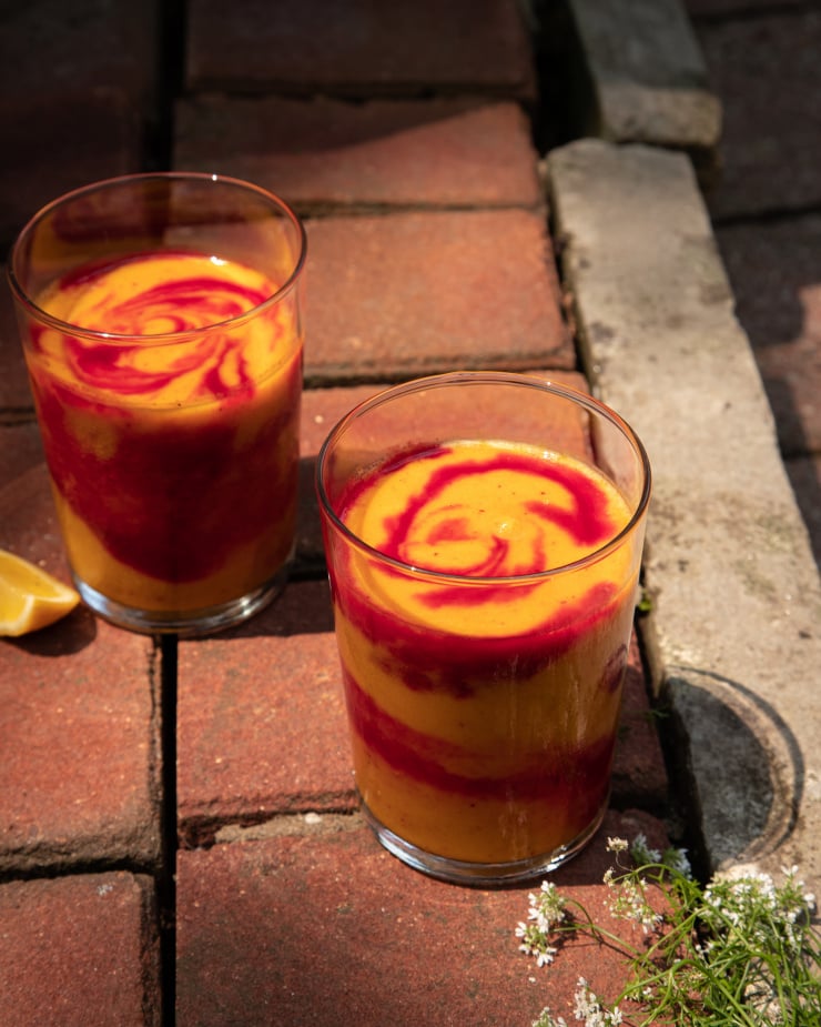 A head on shot of a swirled orange and magenta smoothie in a clear glass. The smoothie is on a red brick background with cilantro flowers nearby.