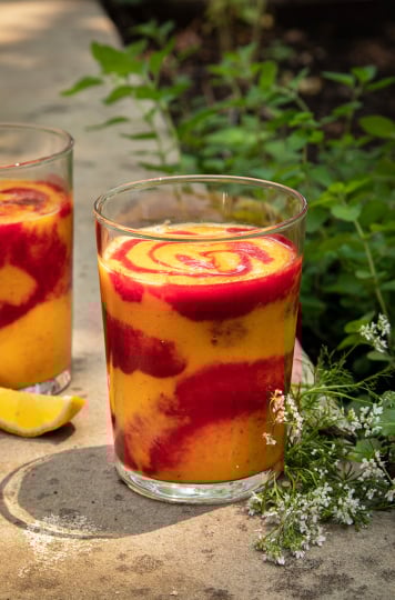 A head on shot of a swirled orange and magenta smoothie in a clear glass. The smoothie is on a concrete background with cilantro flowers nearby.