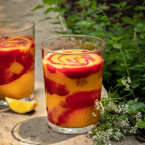 A head on shot of a swirled orange and magenta smoothie in a clear glass. The smoothie is on a concrete background with cilantro flowers nearby.