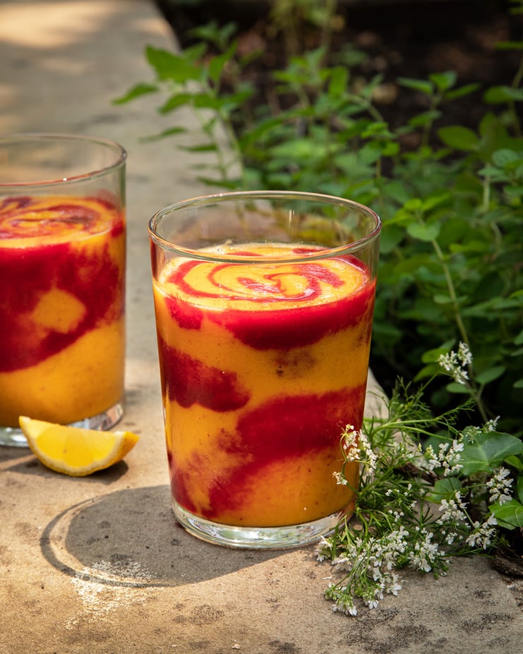 A head on shot of a swirled orange and magenta smoothie in a clear glass. The smoothie is on a concrete background with cilantro flowers nearby.