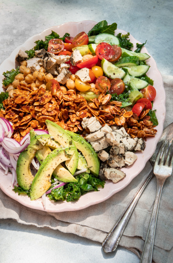 A 3/4 angle shot of a composed salad in bright, outdoor light. Salad is photographed on a link plate and includes: halved cherry tomatoes, kale, sliced cucumber, sliced avocado, chickpeas, toasted almond "bacon", sliced red onion, and bits of crumbled vegan cheese.