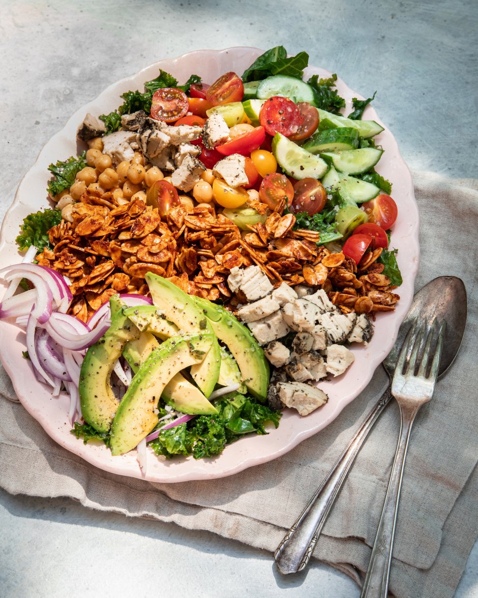 A 3/4 angle shot of a composed salad in bright, outdoor light. Salad is photographed on a link plate and includes: halved cherry tomatoes, kale, sliced cucumber, sliced avocado, chickpeas, toasted almond "bacon", sliced red onion, and bits of crumbled vegan cheese.