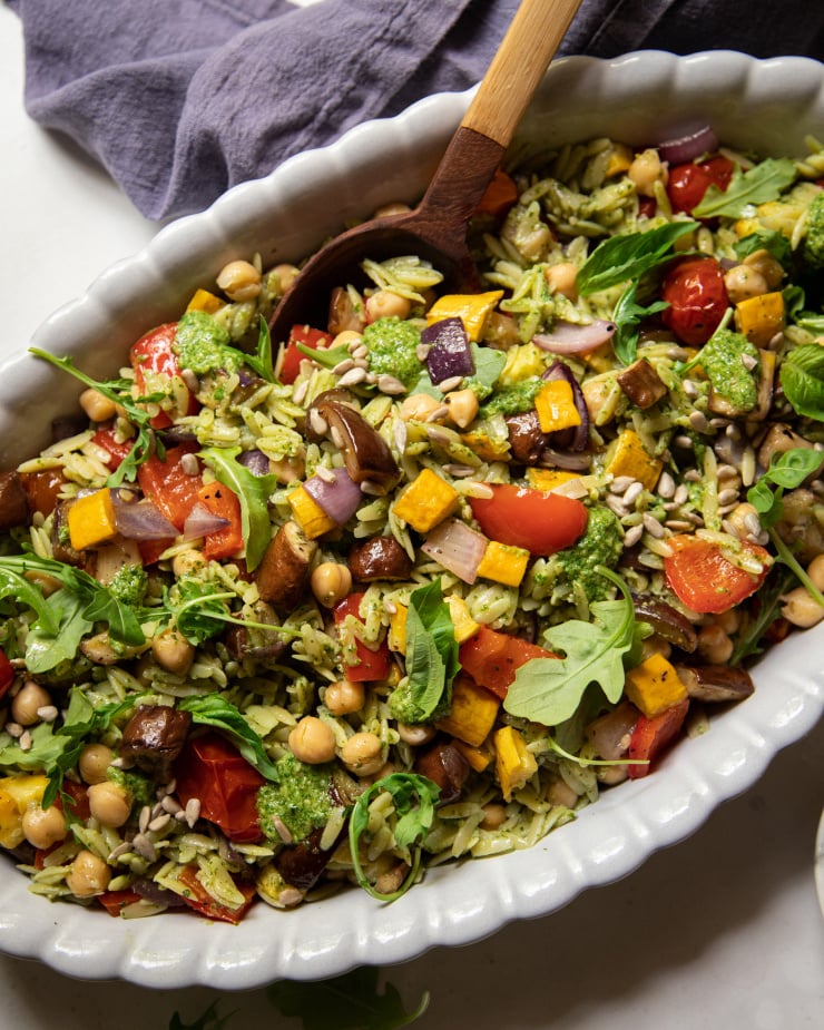 An up close, overhead shot of a pesto orzo salad with dices of roasted vegetables and chickpeas. The dressing is creamy and bright green. There is a wooden spoon sticking out of the dish of salad.