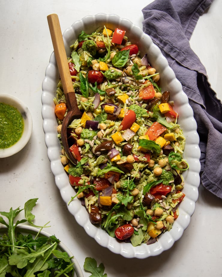 An overhead shot of a pesto orzo salad with dices of roasted vegetables and chickpeas. The dressing is creamy and bright green. There is a wooden spoon sticking out of the dish of salad.