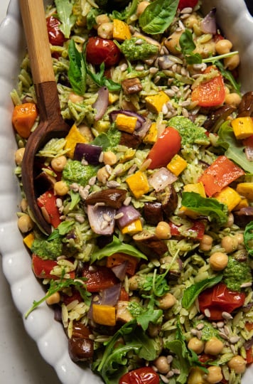 An up close, overhead shot of a pesto orzo salad with dices of roasted vegetables and chickpeas. The dressing is creamy and bright green. There is a wooden spoon sticking out of the dish of salad.