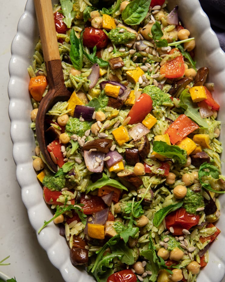 An up close, overhead shot of a pesto orzo salad with dices of roasted vegetables and chickpeas. The dressing is creamy and bright green. There is a wooden spoon sticking out of the dish of salad.