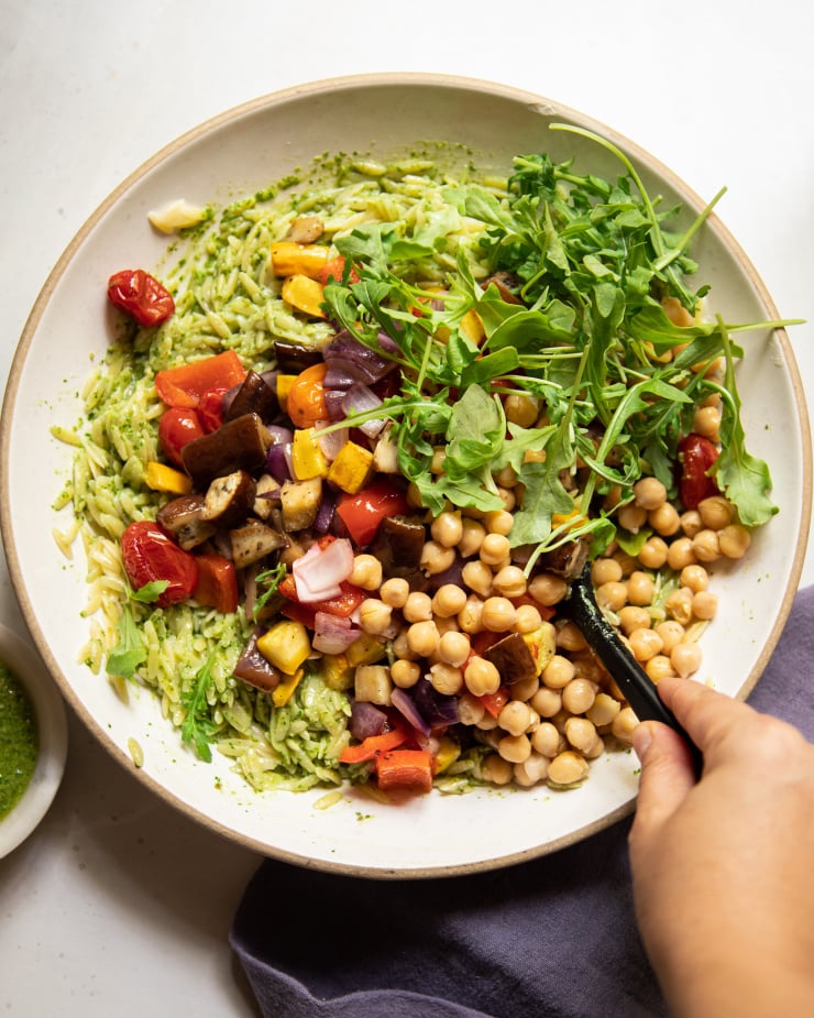 An overhead image shows a hand stirring together an orzo salad with vegetables and chickpeas.