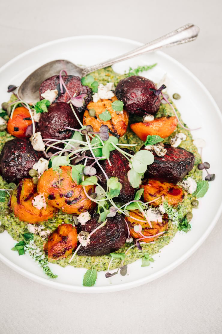 An overhead shot of some grilled beets on top of a textured green sauce on a white plate. The dish is garnished with purple and green radish micro greens and small bits of creamy white vegan cheese.