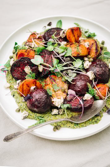 An overhead shot of some grilled beets on top of a textured green sauce on a white plate. The dish is garnished with purple and green radish micro greens and small bits of creamy white vegan cheese.