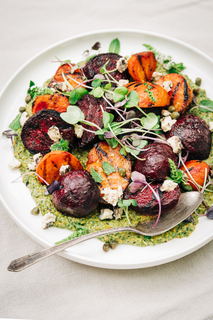 An overhead shot of some grilled beets on top of a textured green sauce on a white plate. The dish is garnished with purple and green radish micro greens and small bits of creamy white vegan cheese.