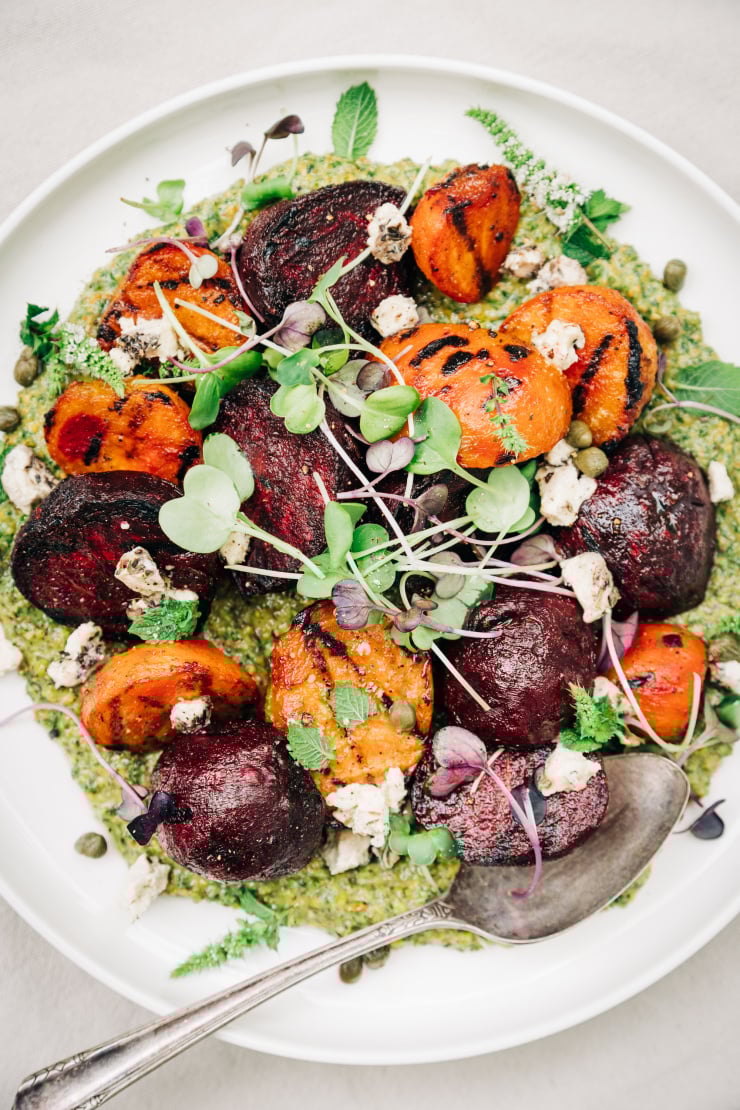An overhead shot of some grilled beets on top of a textured green sauce on a white plate. The dish is garnished with purple and green radish micro greens and small bits of creamy white vegan cheese.