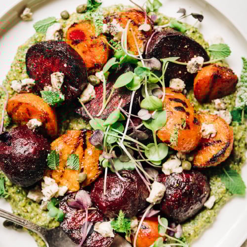 An overhead shot of some grilled beets on top of a textured green sauce on a white plate. The dish is garnished with purple and green radish micro greens and small bits of creamy white vegan cheese.