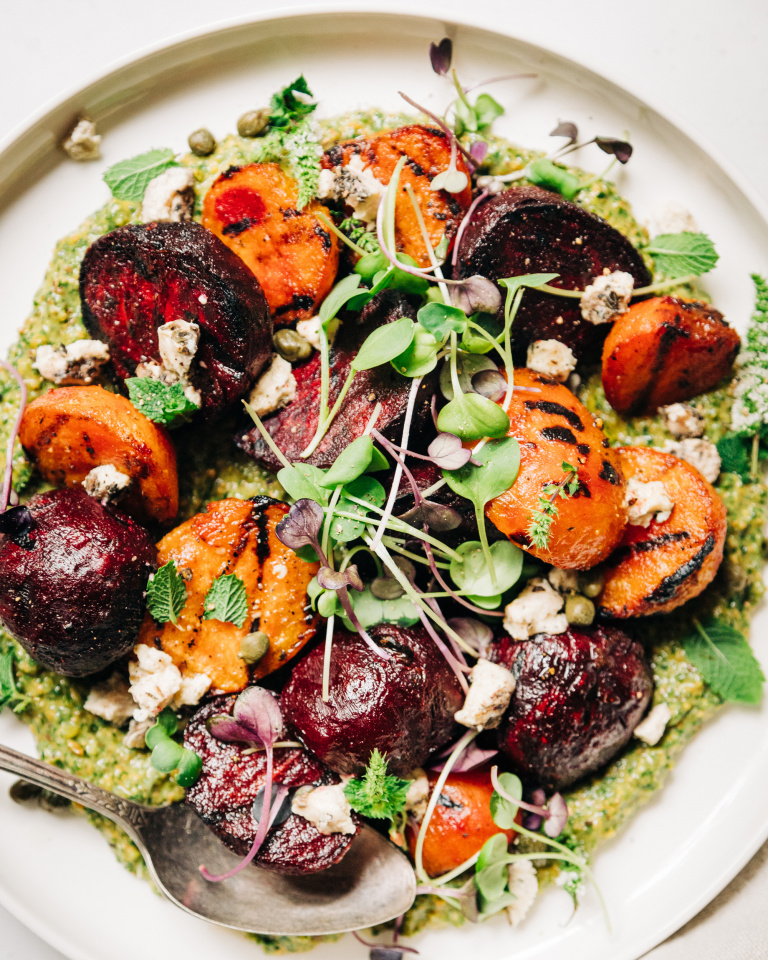 An overhead shot of some grilled beets on top of a textured green sauce on a white plate. The dish is garnished with purple and green radish micro greens and small bits of creamy white vegan cheese.