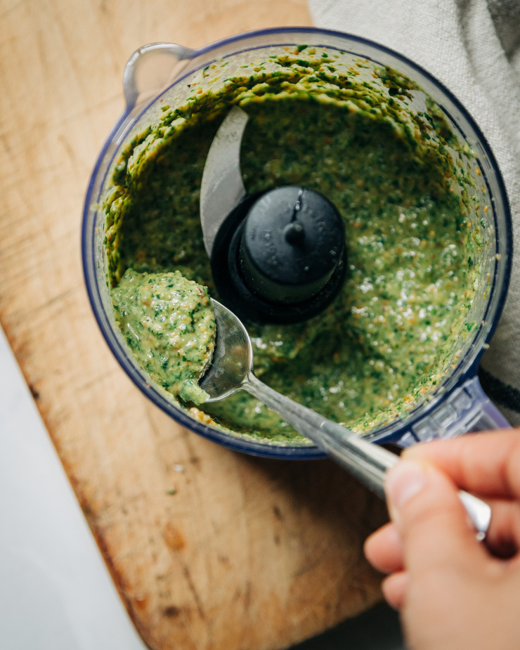 Image shows a hand holding a spoonful of green pesto sauce out of a food processor bowl.