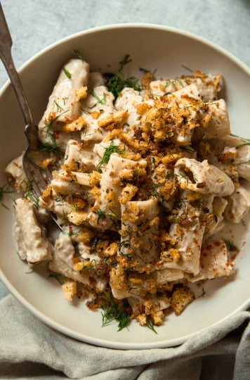 An overhead shot of a bowl of creamy eggplant pasta that is topped with golden, crunchy breadcrumbs, chili flakes, and fresh bits of dill. There is a fork hanging out of the bowl.