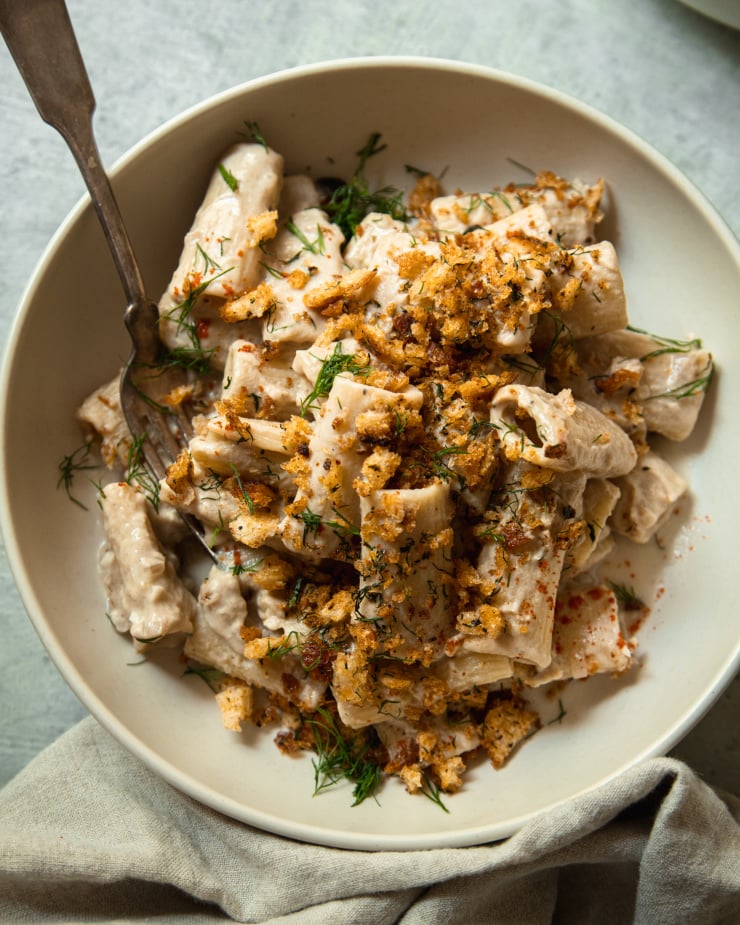 An overhead shot of a bowl of creamy eggplant pasta that is topped with golden, crunchy breadcrumbs, chili flakes, and fresh bits of dill. There is a fork hanging out of the bowl.