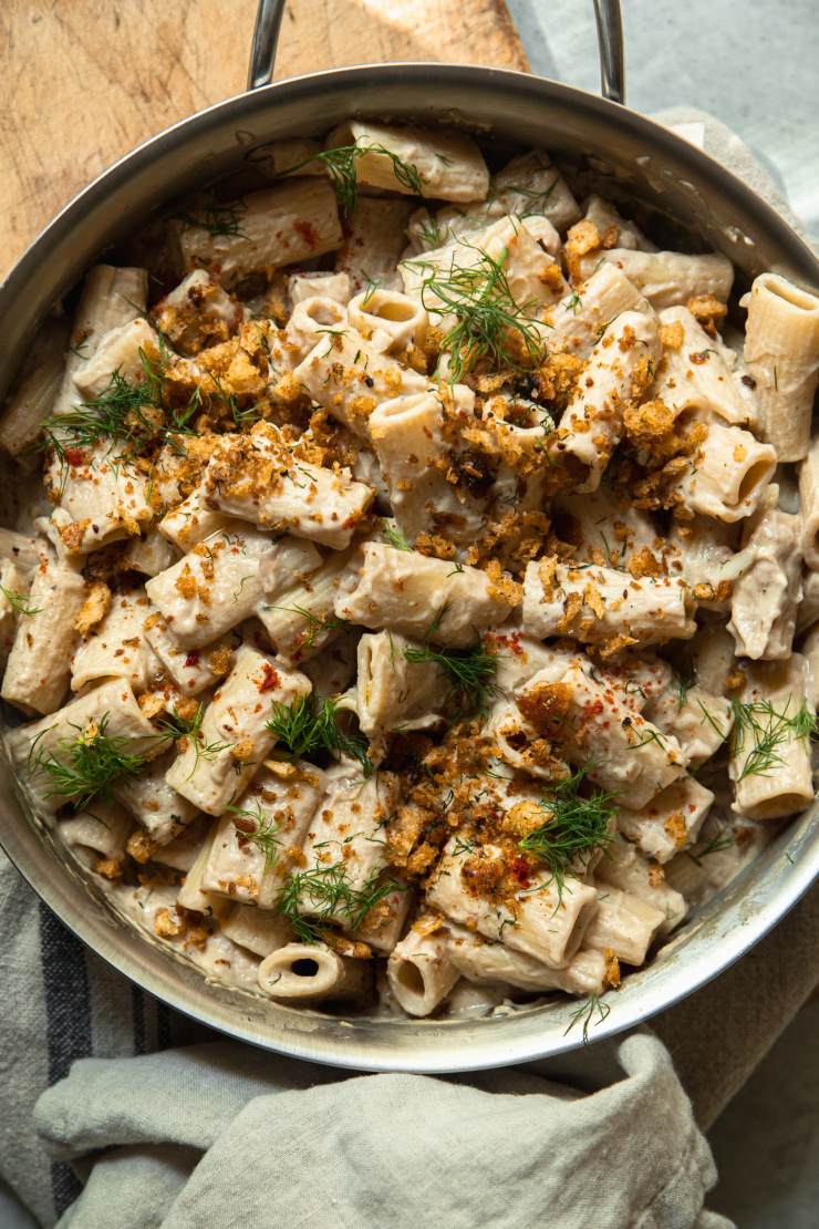 An overhead image of a creamy rigatoni pasta in a stainless steel frying pan. The pasta is topped with golden crunchy breadcrumbs, chili flakes, and chopped bits of fresh dill.