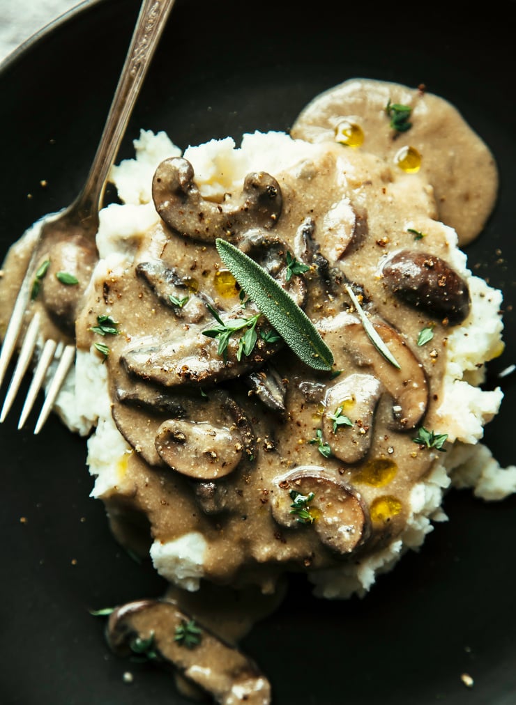 An overhead shot of creamy mushroom gravy that has been spooned over mashed potatoes. The gravy is garnished with whole sage and thyme leaves and ground pepper.