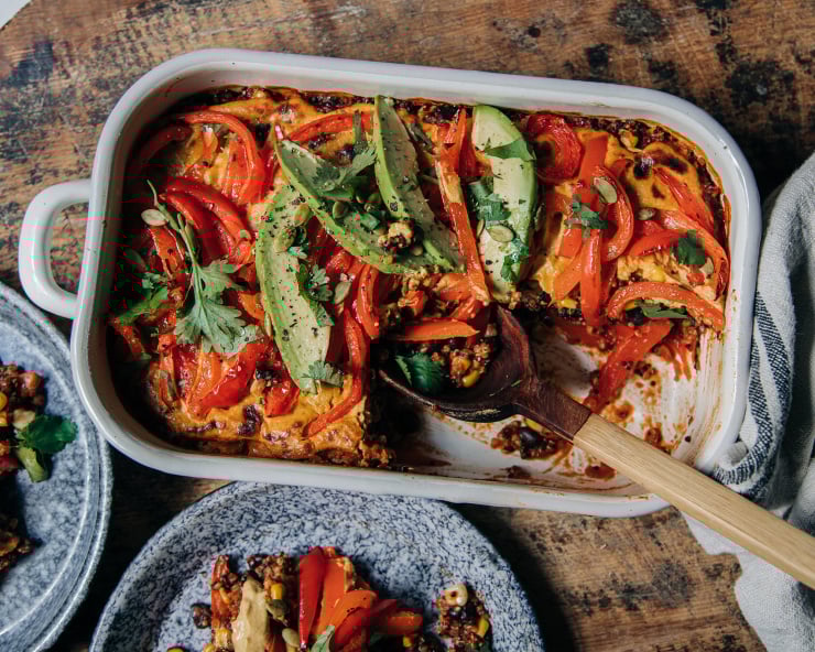 An overhead shot of a vegan casserole topped with sautéed bell peppers, avocado, and chopped cilantro. The casserole dish is white and perched on top of a worn wood surface.