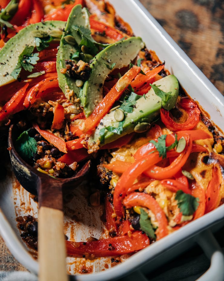 A 3/4 shot of a vegan casserole topped with sautéed bell peppers, avocado, and chopped cilantro. The casserole dish is white and perched on top of a worn wood surface.