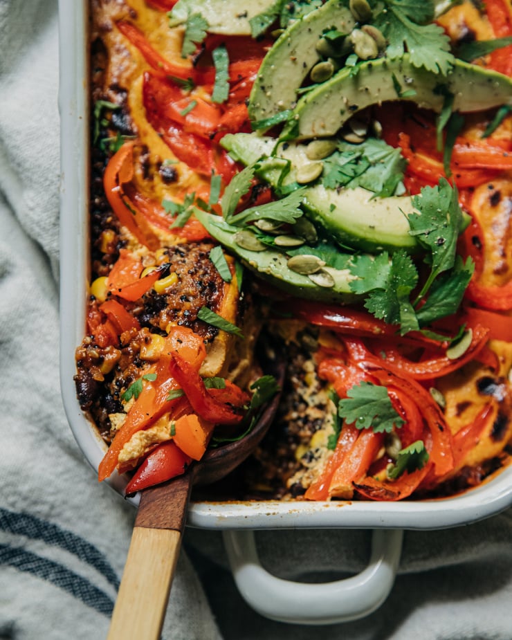 An overhead shot of a vegan stuffed pepper casserole topped with sautéed bell peppers, avocado, and chopped cilantro. The casserole dish is white and a wood serving spoon is taking out a portion of the dish.
