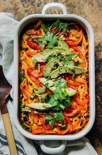 An overhead shot of a vegan casserole topped with sautéed bell peppers, avocado, and chopped cilantro. The casserole dish is white and perched on top of a worn wood surface.
