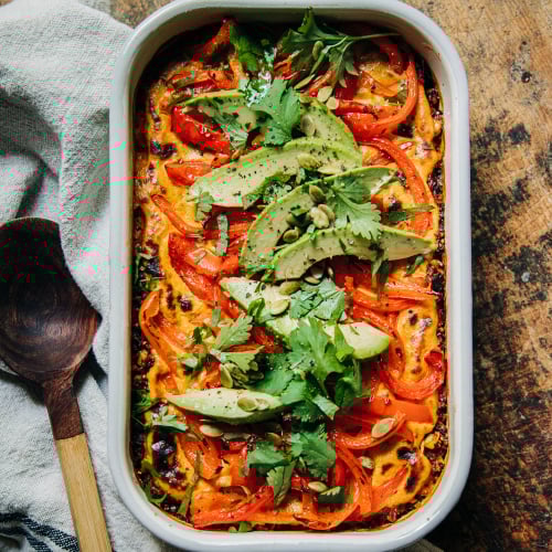 An overhead shot of a vegan casserole topped with sautéed bell peppers, avocado, and chopped cilantro. The casserole dish is white and perched on top of a worn wood surface.