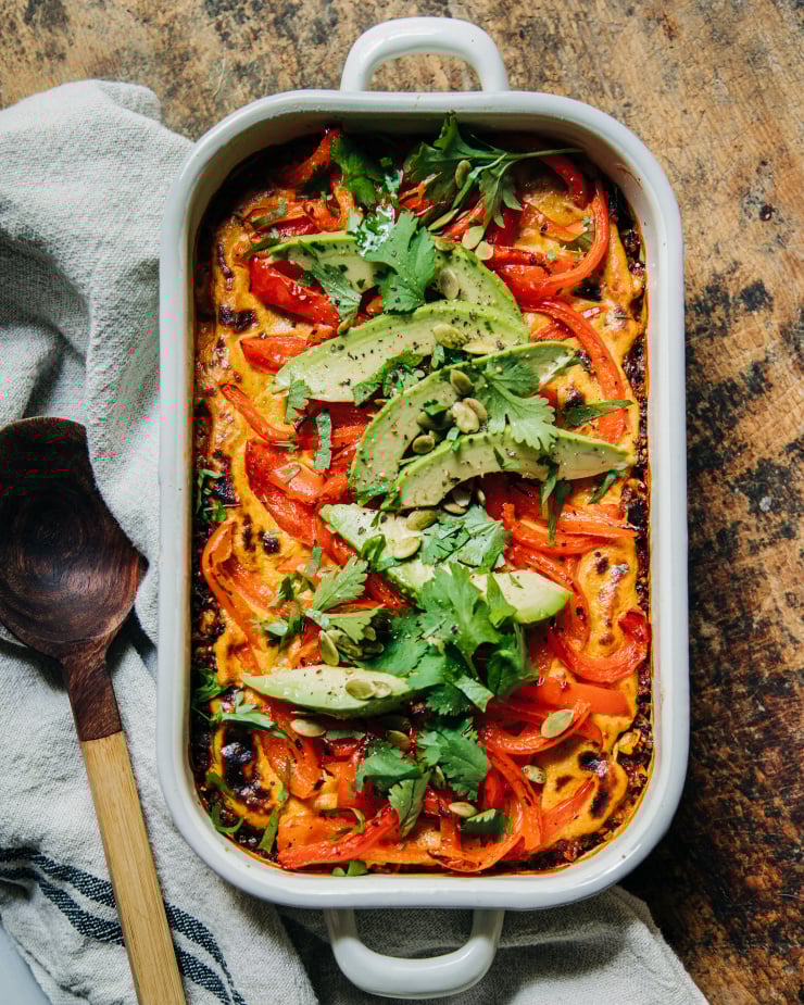 An overhead shot of a vegan stuffed pepper casserole topped with sautéed bell peppers, avocado, and chopped cilantro. The casserole dish is white and perched on top of a worn wood surface.