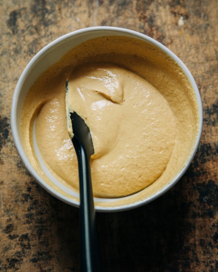 An overhead shot of a creamy, pale orange, vegan "cheese" sauce in a white bowl on a worn wood background.