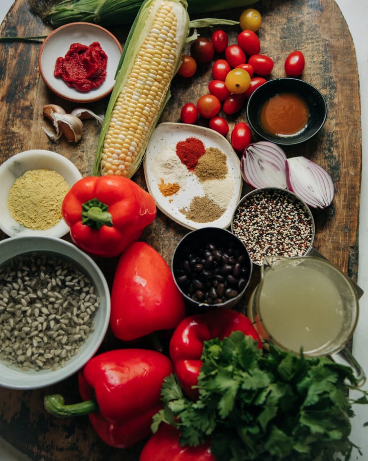 An overhead image of ingredients for a hearty vegan meal, including corn on the cob, a can of black beans, various spices, red bell peppers, cilantro, cherry tomatoes, garlic, and quinoa.